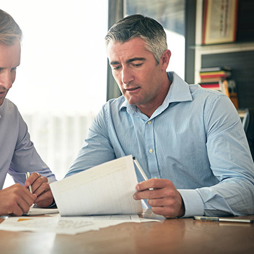 twee mannen in een lichtblauw overhemd zitten aan een tafel. De oudere man op rechts wijst naar een papier wat hij de jongere man op links laat zien. Er staat een koffiekopje op tafel.